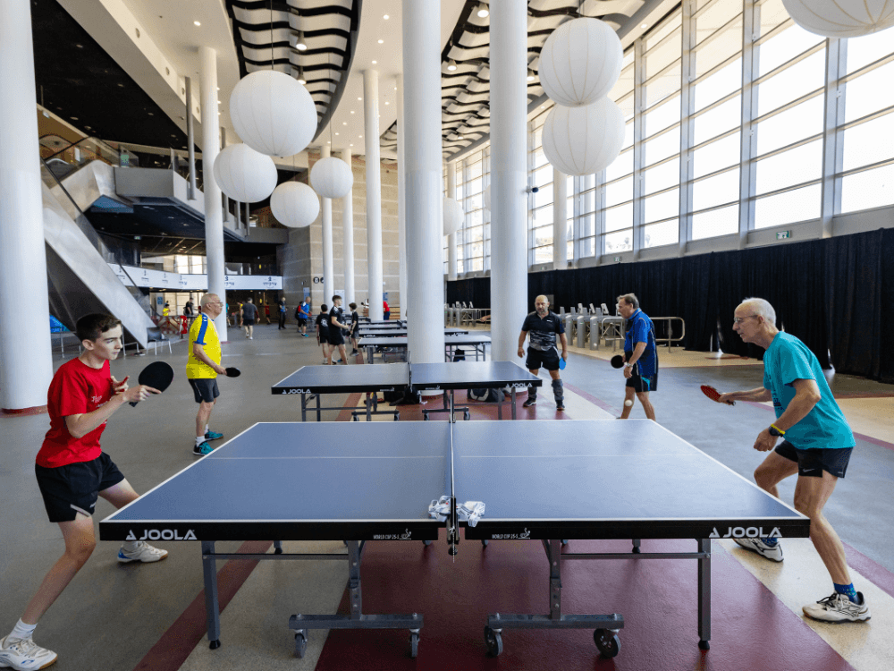 Atletas de diferentes idades jogam tênis de mesa em várias mesas instaladas em um amplo espaço destinado à prática esportiva.