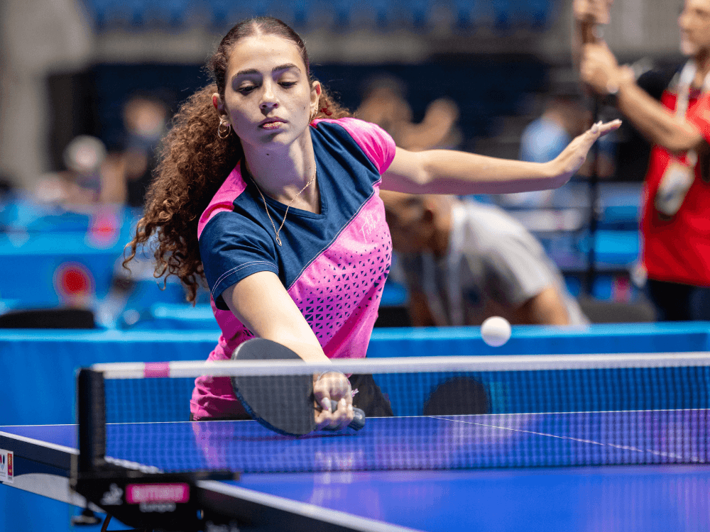 Jogadora de tênis de mesa com uniforme rosa e azul rebate a bola durante uma partida competitiva em uma mesa azul.